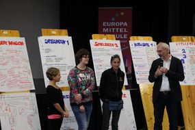 Drei Kinder und ein Mann stehen vor den zuvor gestalteten Plakaten bei der Spielforscher-Werkstatt in der Europa-Mittelschule Prinzersdorf.