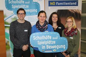 Ein Mann und drei Frauen halten ein blaues Schild mit "Schulhöfe und Spielplätze in Bewegung" in die Kamera.