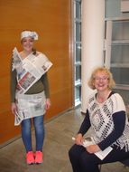 Ein Mädchen trägt einen Hut, ein T-Shirt und einen Rock aus Zeitungspapier über ihrem Gewand und lächelt zusammen mit einer Dame in die Kamera