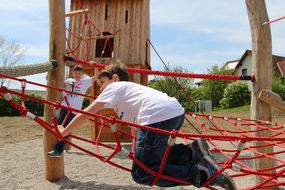 Zwei Buben klettern auf roten Seilen zum Holzhaus hinauf.
