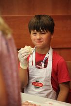Bub in rotem T-Shirt isst bei der NÖ Kinder Business Week ein Sandwich und lächelt glücklich in die Kamera