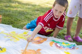 Ein Bub bemalt ein weißes, großes Tuch mit bunten Farben