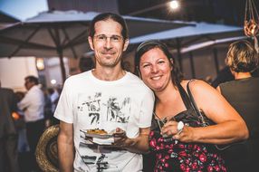 Ein Mann und eine Frau lachen mit Speisen und Getränken in der Hand in die Kamera.